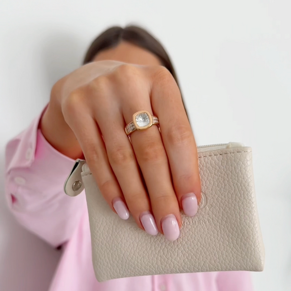 Hand holding a small beige pouch with a gold ring on a plain background